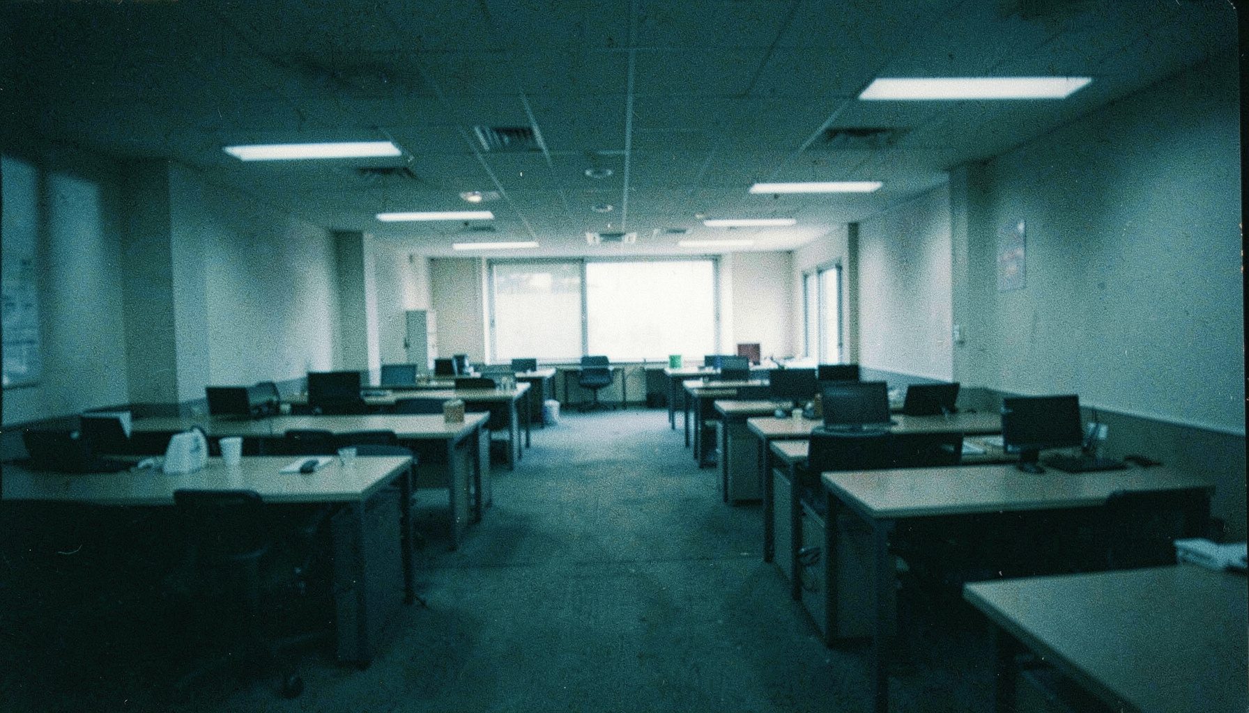 A wide dim office workspace photographed on grainy analog film, deep teal and charcoal tones, rows of empty desks and soft overhead light suggesting a quiet shared work environment